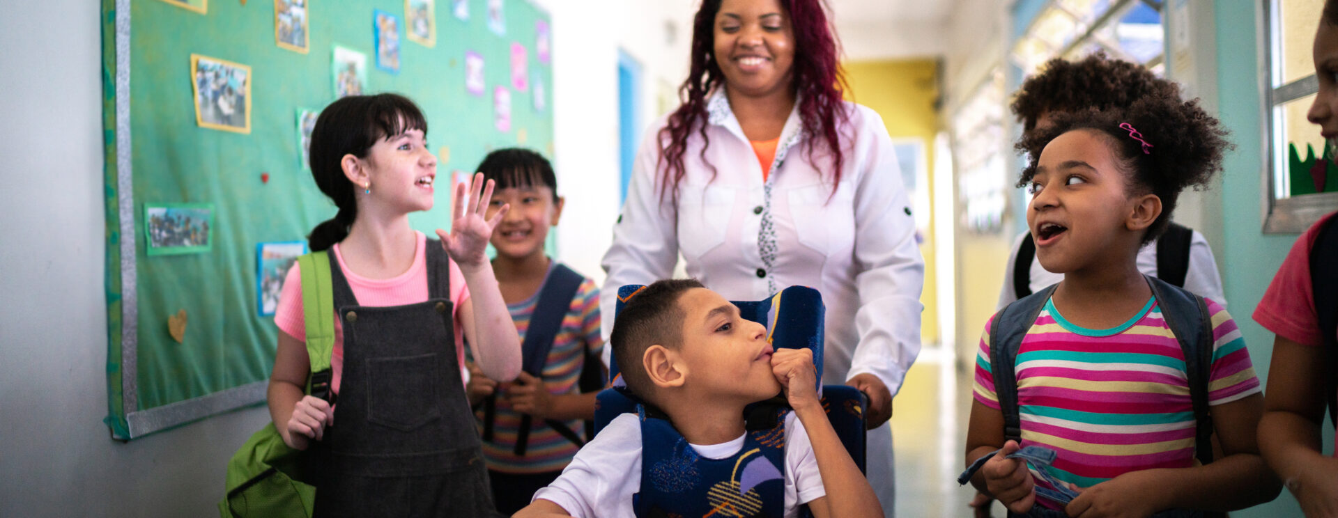 Groupe d'élèves dans un couloir d'école. Au premier plan, un élève en fauteuil, poussé par une femme adulte. Tous sont souriants.