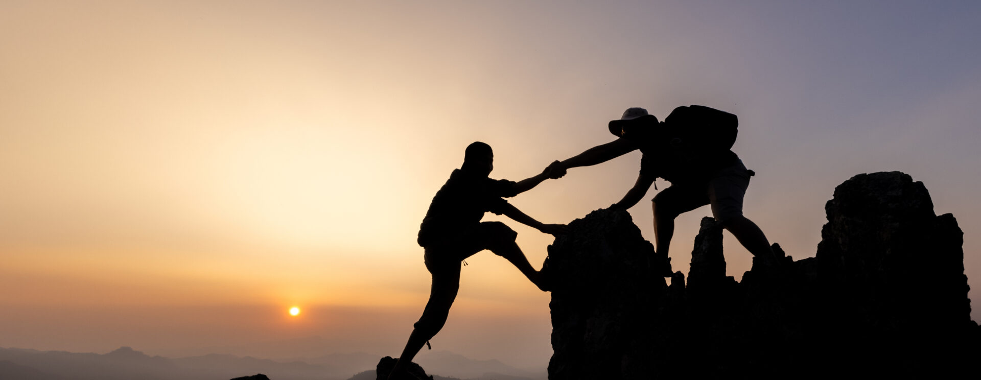 Un paysage de montagne au soleil couchant. On voit la silouheute de deux randonneurs : l'un, au sommet, aide l'autre à franchir le dernier pas qui est difficile.