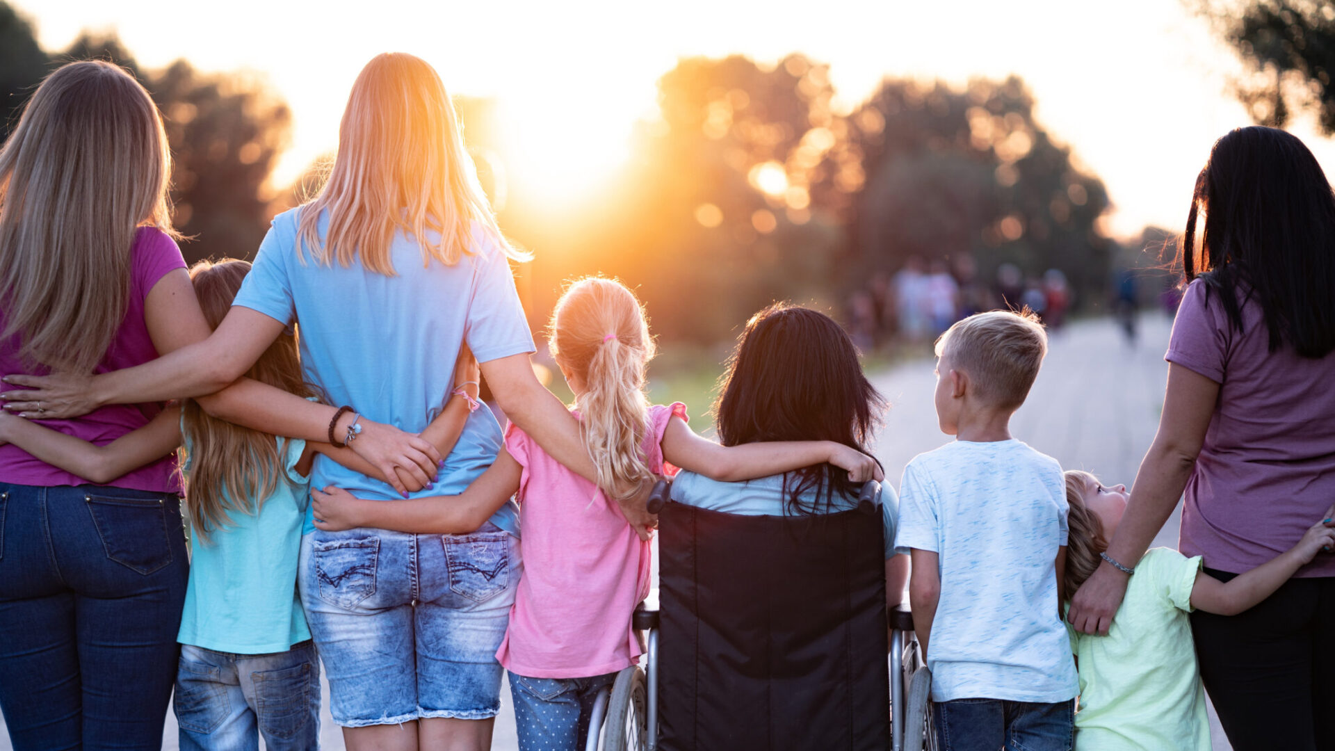 Des enfants, dont une en fauteuil, et trois adultes, se tiennent par les épaules. Ils sont de dos, à l'extérieur, habillés comme en été et le soleil est couchant.